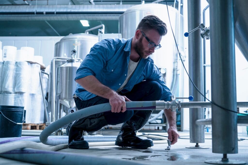 Young man in brewery crouching down connecting pipe